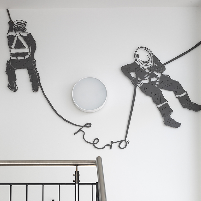 Kunst am Bau: An der weißen Wand im Treppenhaus hängt ein Kunstwerk das 2 Feuerwehrmitarbeiter:innen zeigt die an einem Schlauch hängen, der Schlauch formt das englische Wort "hero"