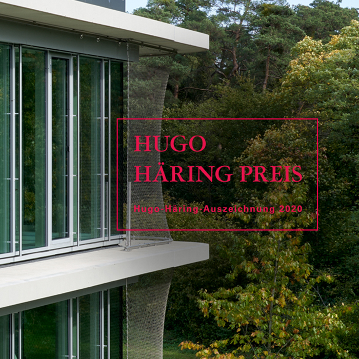 Das Logo des Hugo Häring Preises in grellem Rot vor einer klassischen Glasfassade und Bäumen